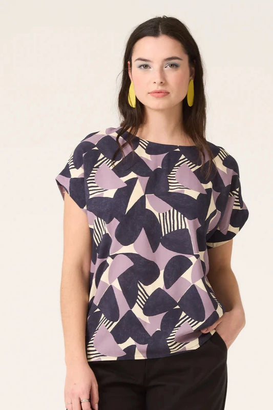 3/4 shot of a woman wearing the Ombre Top by Cokluch in Lilac Half Moon Print, featuring a round neck, short dropped sleeves, and a loose fit in OEKO-TEX certified viscose, with dark pants, standing in front of white background 