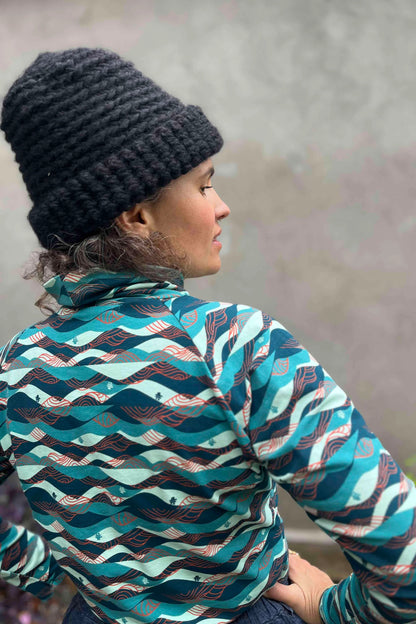 A woman wearing the Oris Raglan Turtleneck by Kazak in Inuksuk print, with a pattern of rolling waves and tiny inukshuks in shades of blue. 