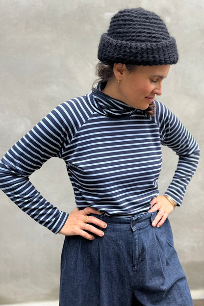 A woman wearing the Oris Raglan Turtleneck by Kazak in Blue Stripe. She is wearing it with jeans and standing outside. 