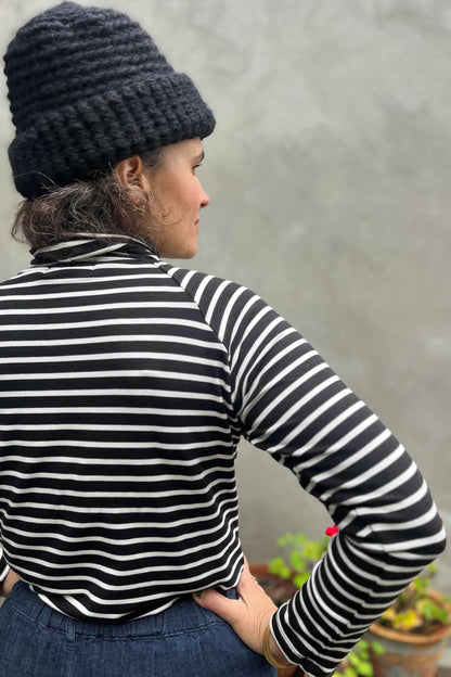 Back view of a woman wearing the Oris Raglan Turtleneck by Kazak in Black Stripe. She is wearing it with jeans and standing outside. 