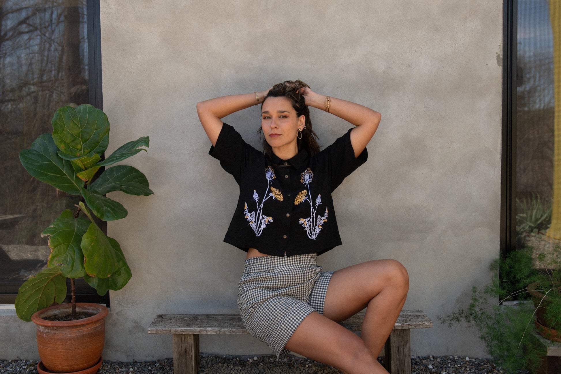 A woman wearing the Hosta Screen-printed Blouse by Kazak in Black with Dandelions, with hand-screen-printed flowers, mid-length raglan sleeves, a classic collar, and a button front. She is wearing it with gingham shorts and sitting outside by a grey wall with plants beside her.