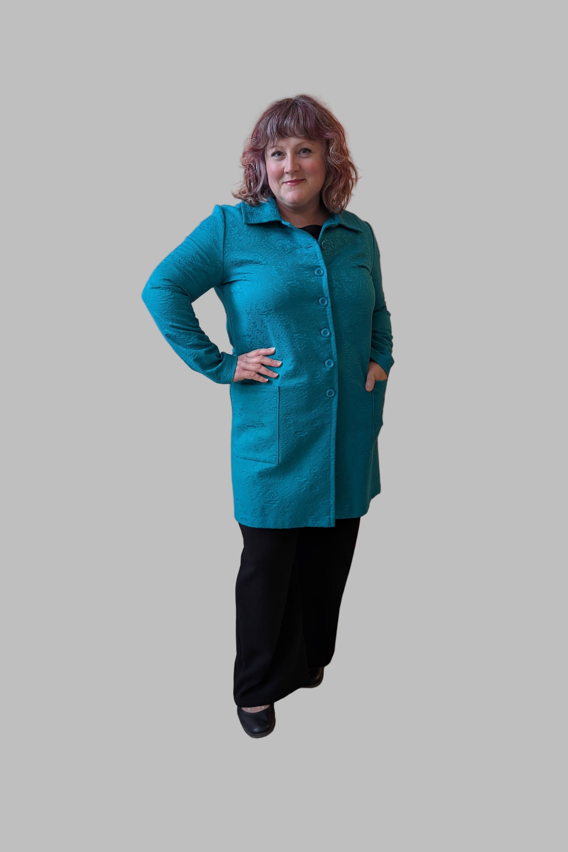 A woman wearing the Emmanuelle Coat by Kollontai in Turquoise jacquard with Black pants. She is standing in front of a white background. 