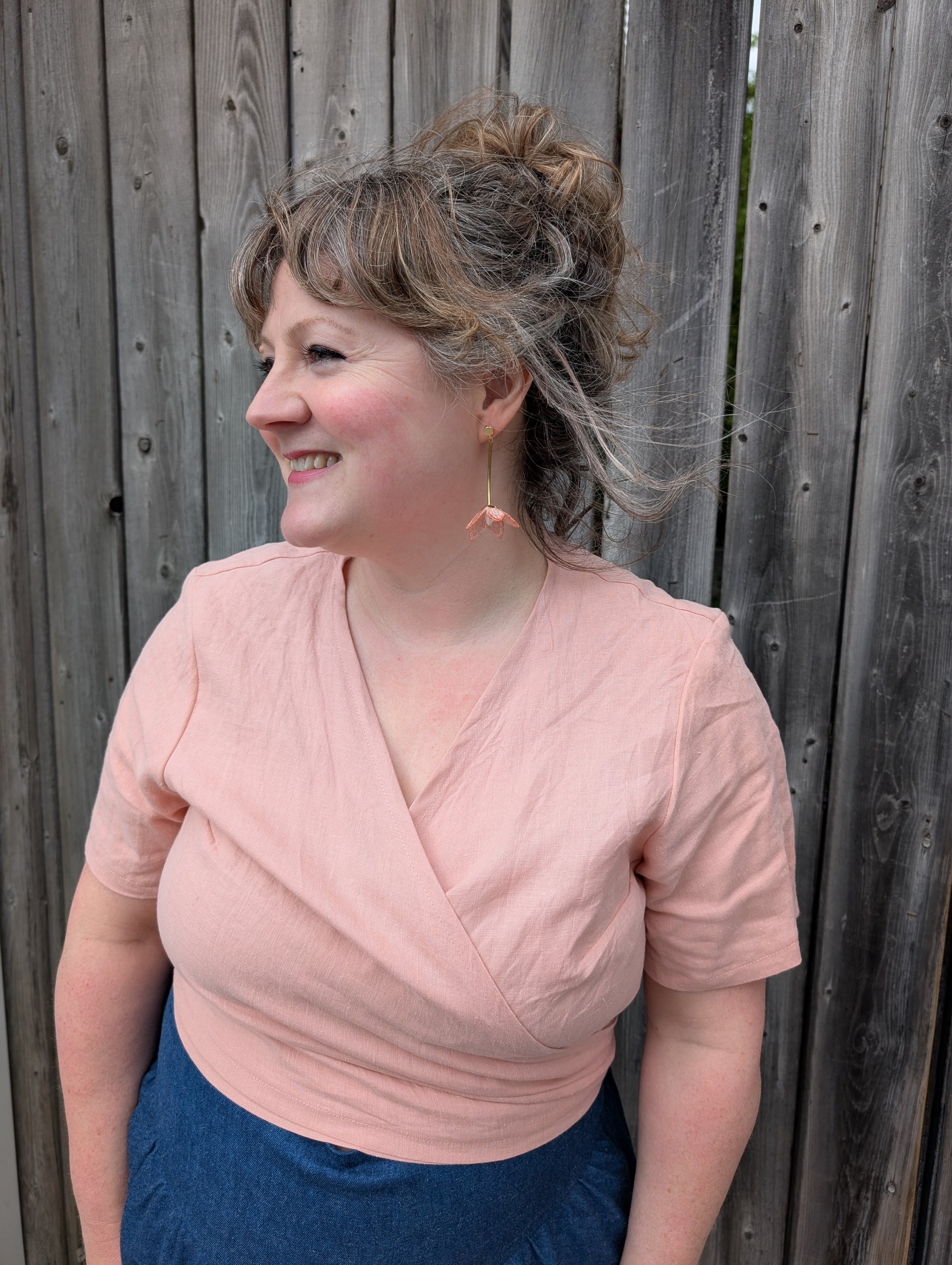 A woman wearing the Stella Top by Lights of All in Ballet Pink, a short sleeved linen wrap top that can be twisted or tied back or front. She is wearing it with the Margot Denim Skirt by Melow and standing by a fence. 