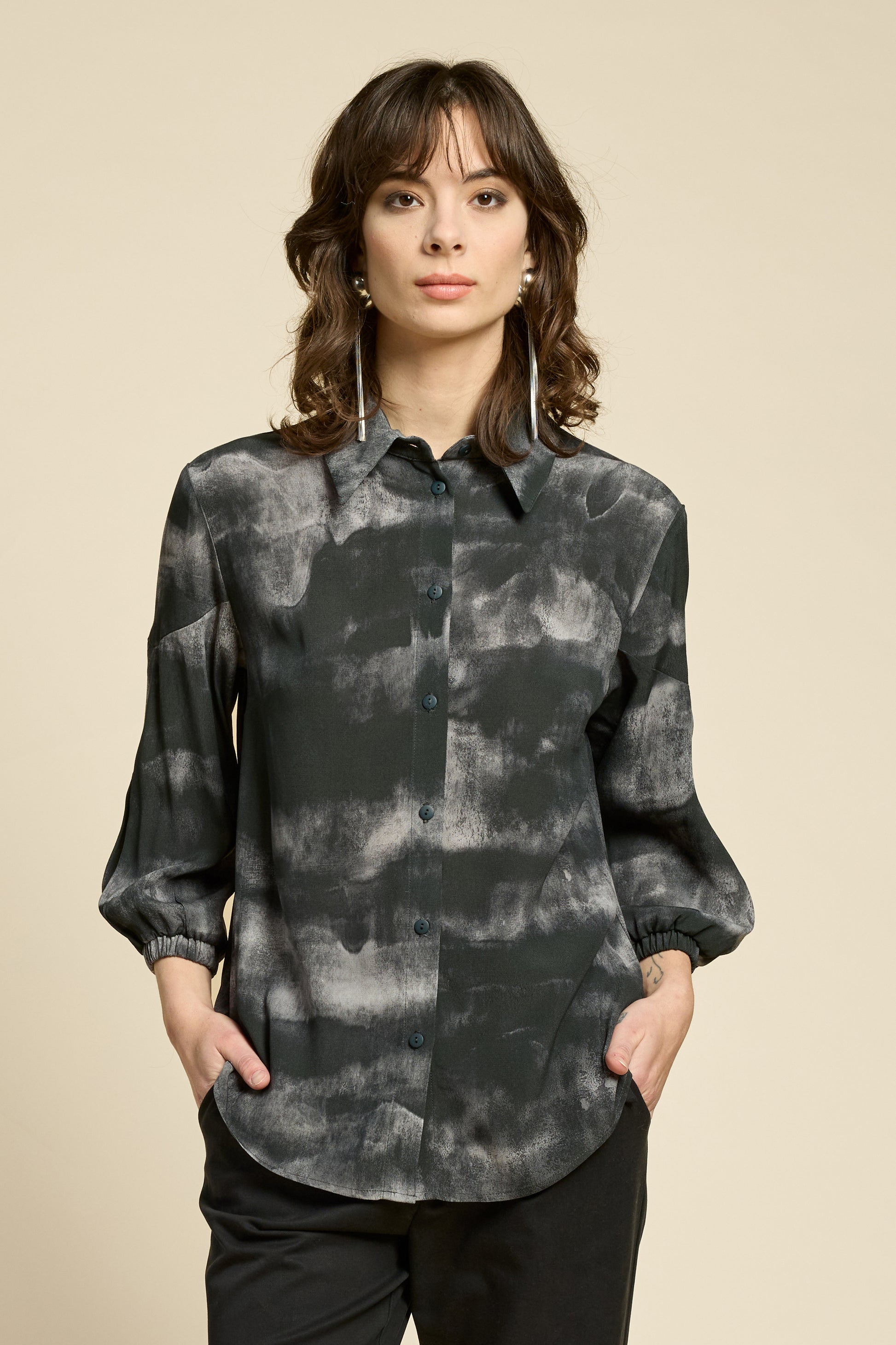 Waist up view of a woman wearing the Petit Chat Blouse by Cokluch in Mist Black, collared button front shirt with slighty puffed 3/4 sleeves and a rounded hem. She is wearing it with black pants and standing in front of a beige background. 
