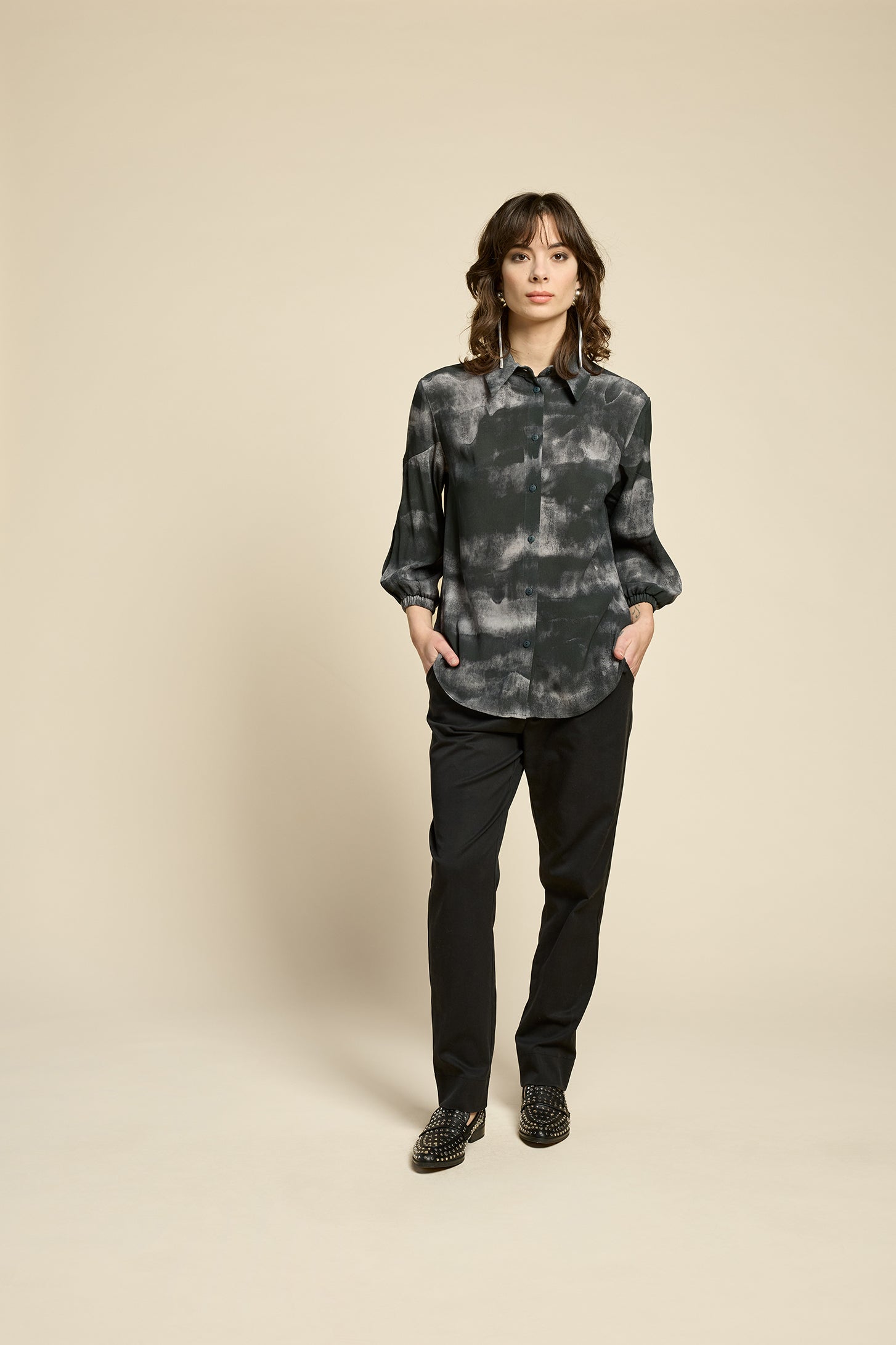 A woman wearing the Petit Chat Blouse by Cokluch in Mist Black, collared button front shirt with slighty puffed 3/4 sleeves and a rounded hem. She is wearing it with black pants and standing in front of a beige background. 
