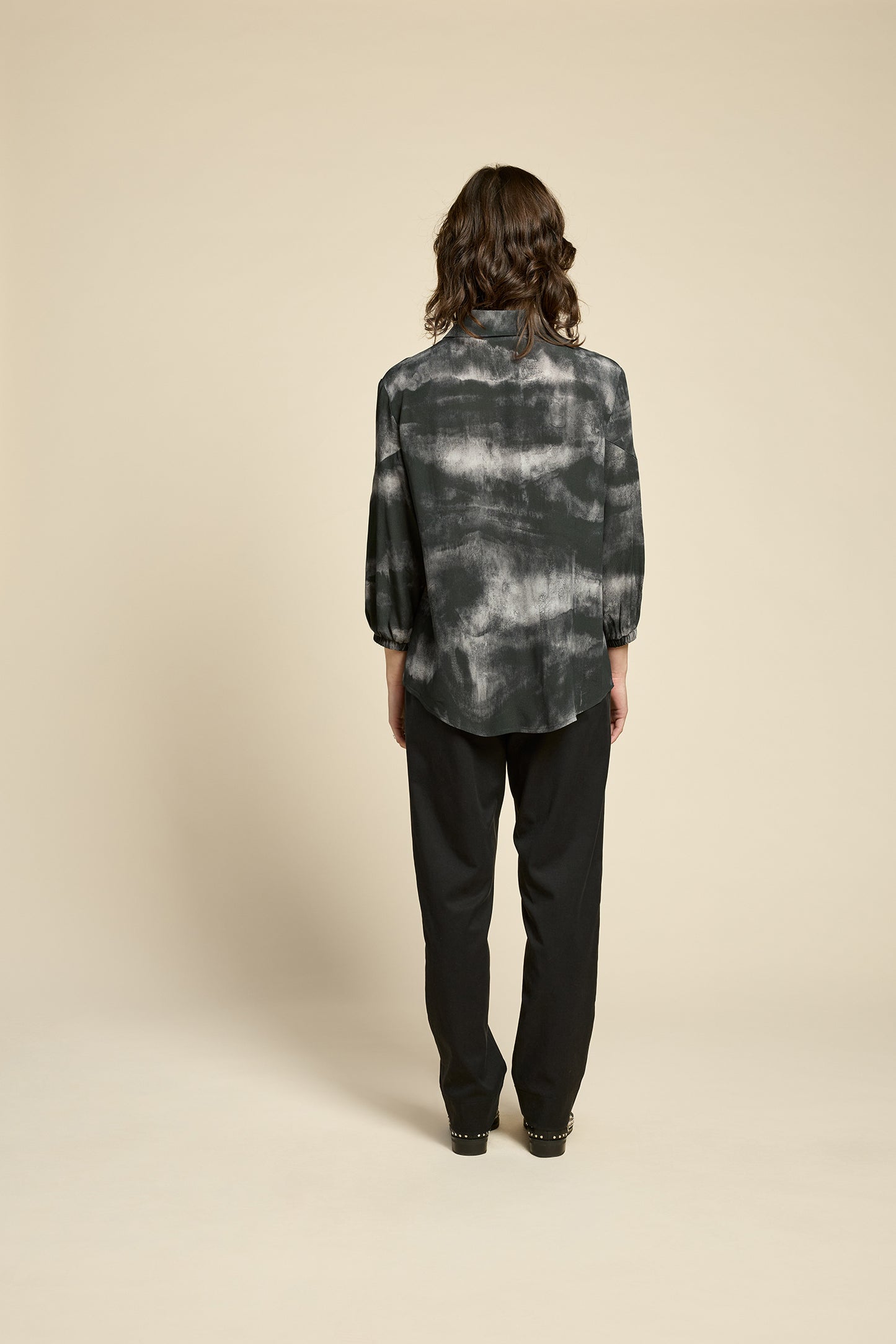 Back view of a woman wearing the Petit Chat Blouse by Cokluch in Mist Black, collared button front shirt with slighty puffed 3/4 sleeves and a rounded hem. She is wearing it with black pants and standing in front of a beige background. 
