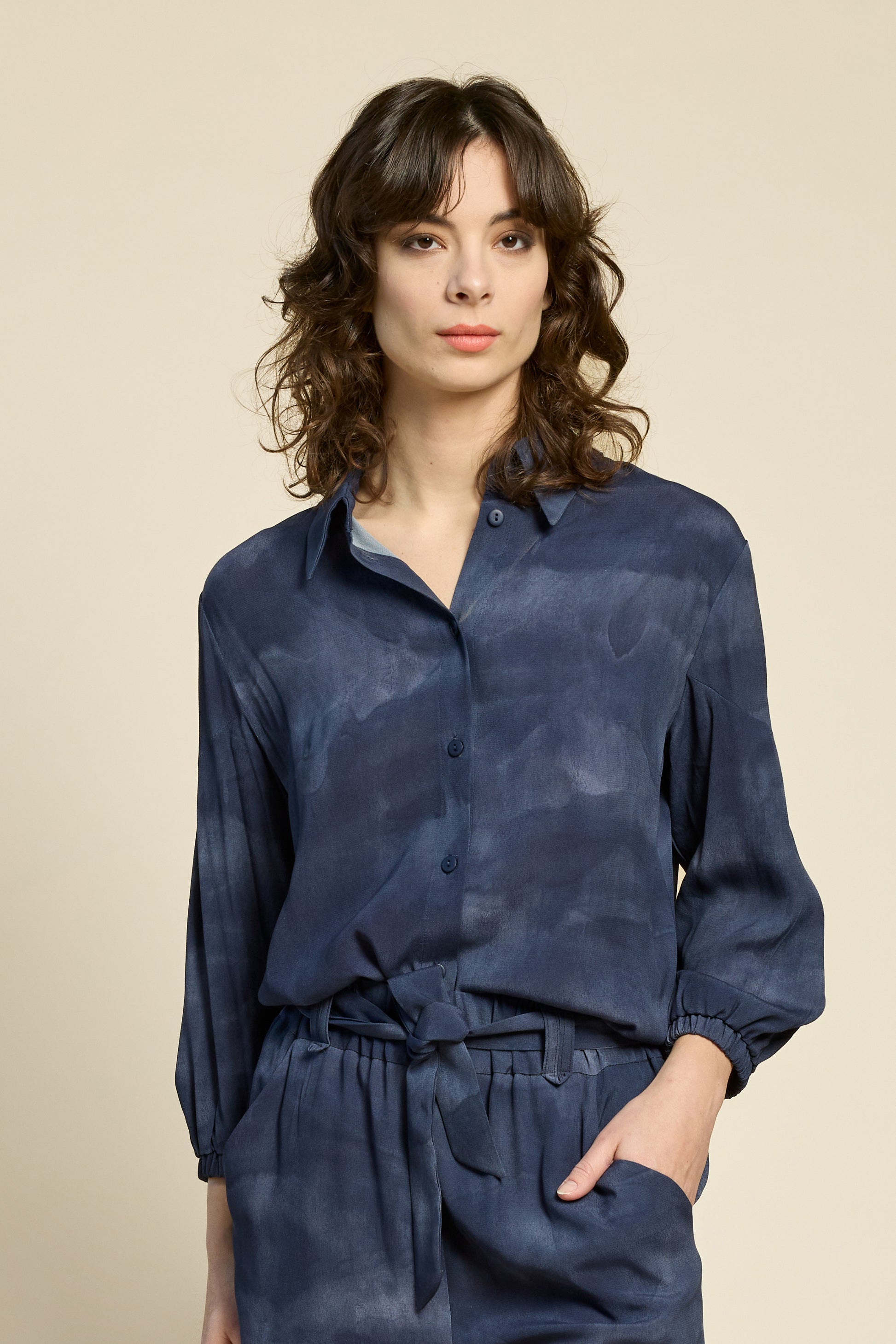 Waist up view of a woman wearing the Petit Chat Blouse by Cokluch in Mist Twilight, a collared button front shirt with slighty puffed 3/4 sleeves and a rounded hem. She is wearing it with matching pants and standing in front of a beige background. 
