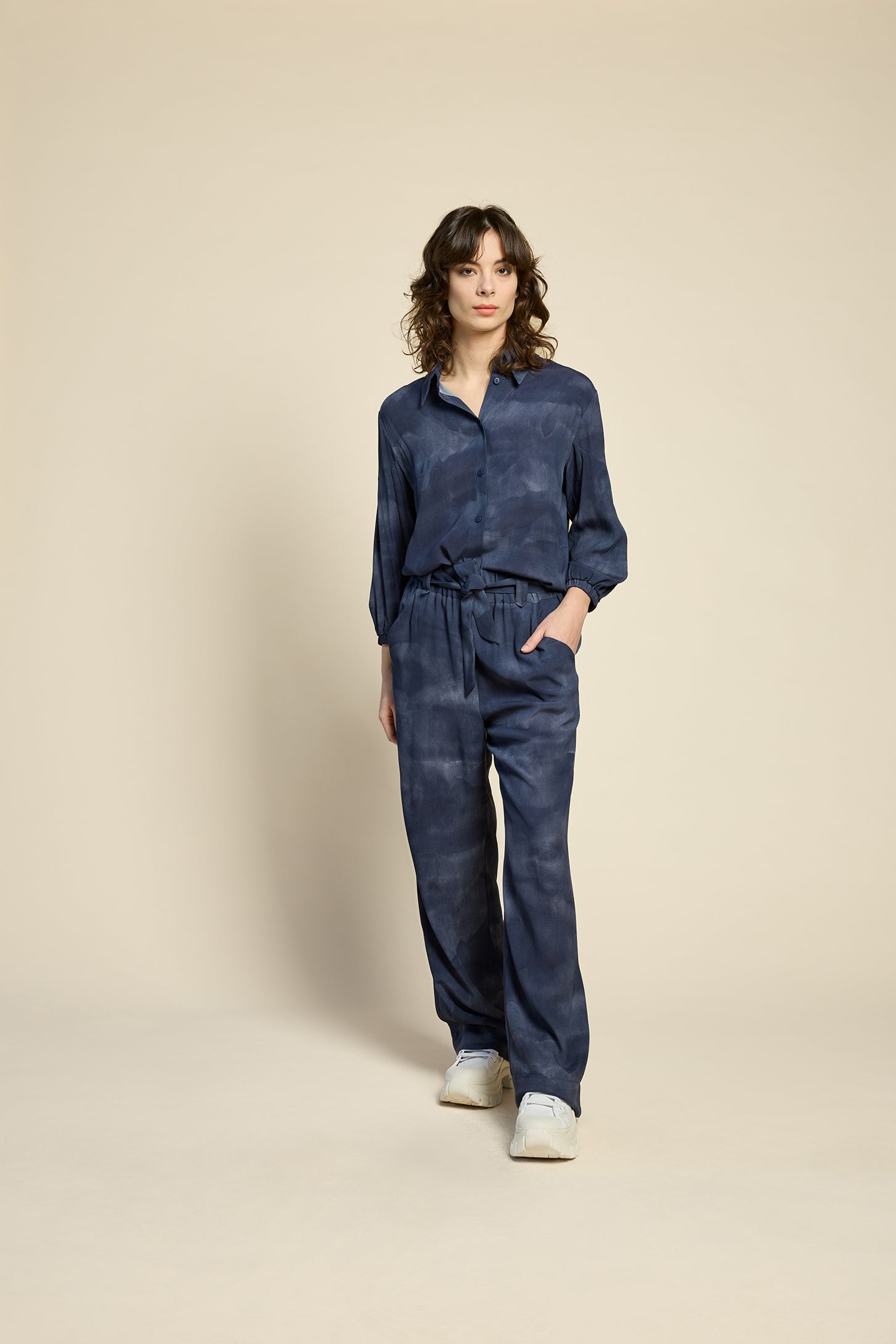 Waist up view of a woman wearing the Petit Chat Blouse by Cokluch in Mist Twilight, a collared button front shirt with slighty puffed 3/4 sleeves and a rounded hem. She is wearing it with matching pants and standing in front of a beige background. 
