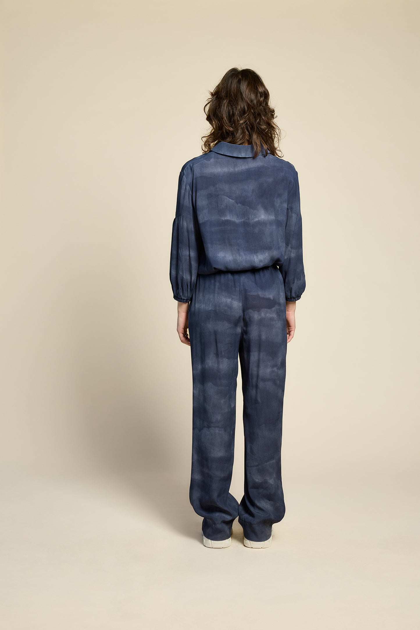 Back view of a woman wearing the Petit Chat Blouse by Cokluch in Mist Twilight, a collared button front shirt with slighty puffed 3/4 sleeves and a rounded hem. She is wearing it with matching pants and standing in front of a beige background. 
