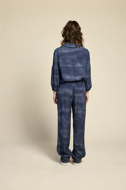 Back view of a woman wearing the Petit Chat Blouse by Cokluch in Mist Twilight, a collared button front shirt with slighty puffed 3/4 sleeves and a rounded hem. She is wearing it with matching pants and standing in front of a beige background. 
