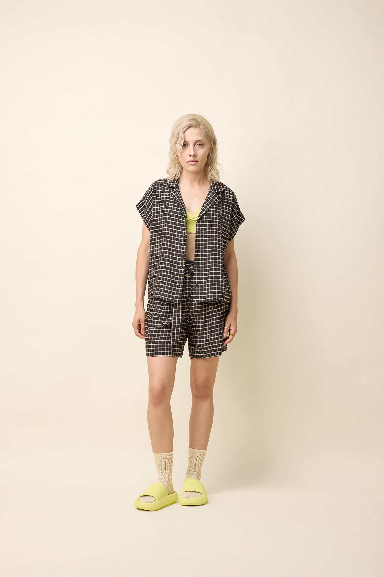 A woman wearing the Phebe Top by Cokluch in Black Utopia, a button front short-sleeved shirt with a rounded blouse collar and a chest pocket. She is wearing it with matching shorts and standing in front of a beige background. 