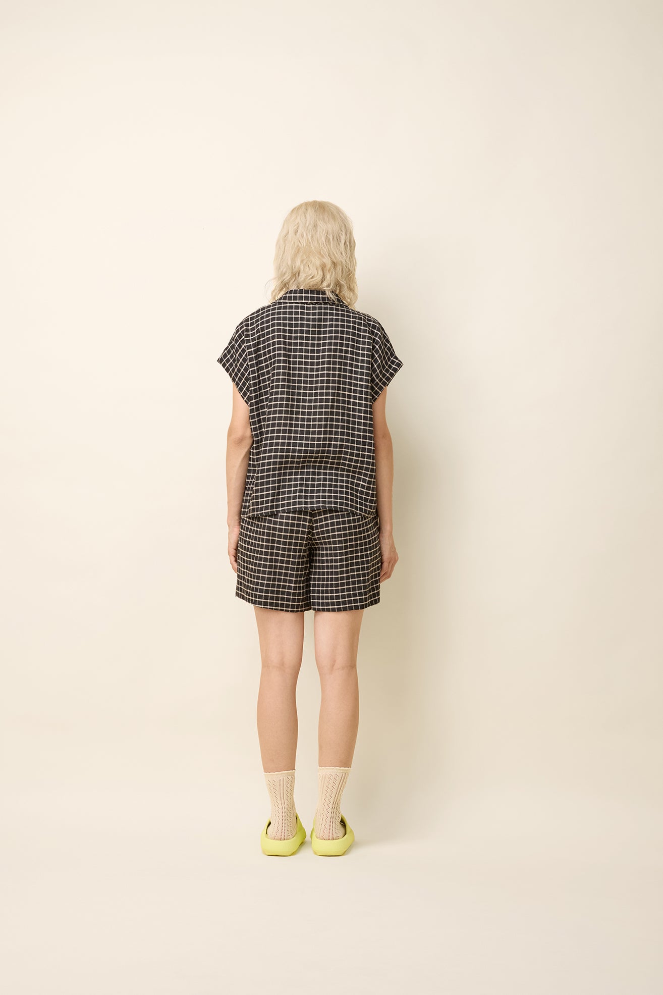 Back view of a woman wearing the Phebe Top by Cokluch in Black Utopia, a button front short-sleeved shirt with a rounded blouse collar and a chest pocket. She is wearing it with matching shorts and standing in front of a beige background. 