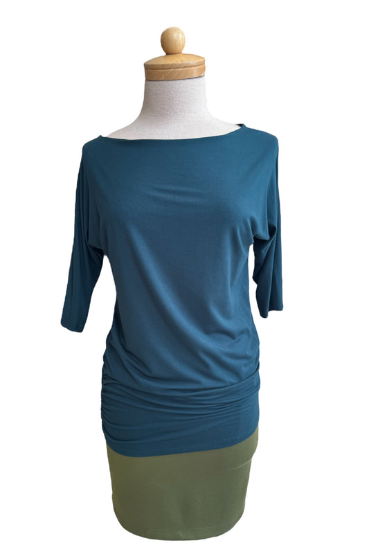 The Bettina Top by MOOVment in Teal, with boat neck, 3/4 sleeves, and gathered sides, is shown with a green skirt on a mannequin in front of a white background. 