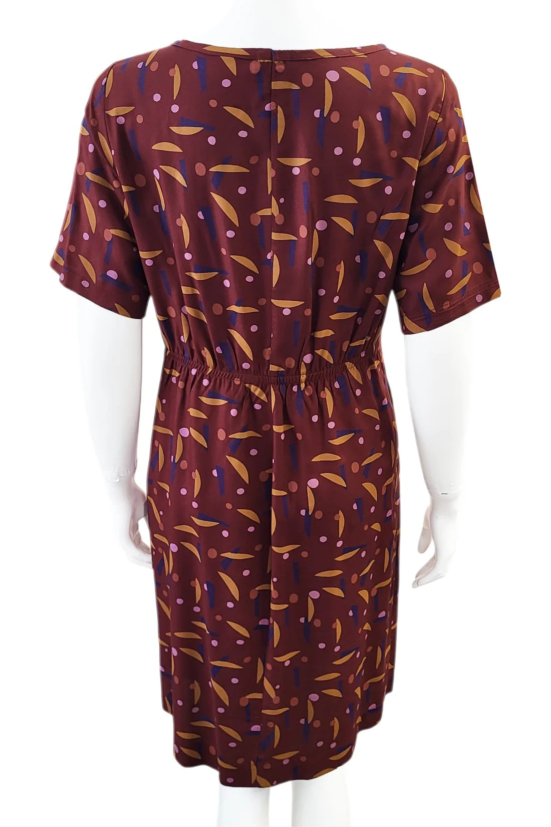 A back view of the Cecily Simple Shift Dress from Mandala in Playtime Rust print is shown on a mannequin against a white background. It has a high neck, short sleeves, a knee-length hem, French darts, and elastic at the back of the waist. 