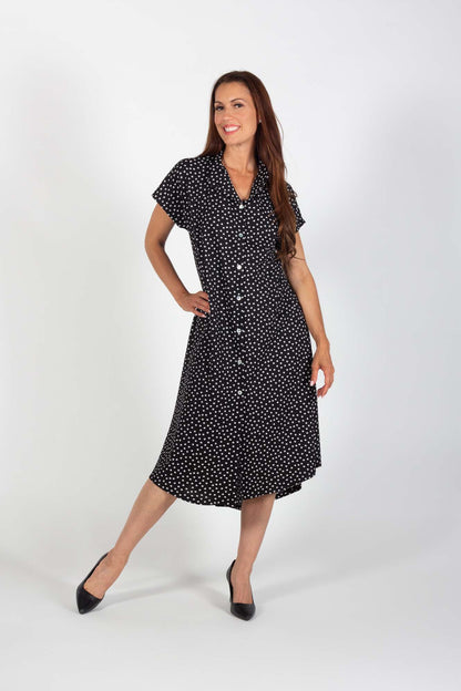 A woman wearing the Cora Dress by Pure Essence in Black with polka dots, standing in front of a coral wall