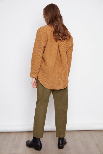 Back view of a woman wearing the Charlie Top by Julie in Ginger Stripes, with an asymmetrical button placket at the neckline and hi-low hemline with side slits. She's wearing them with green pants and standing in front of a white background. 