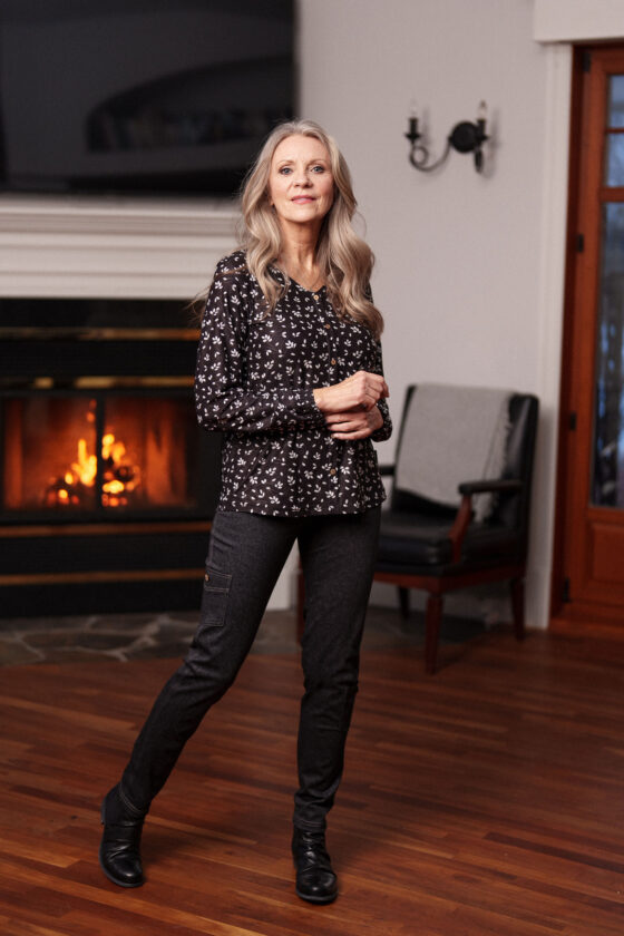 A woman wearing the Monet Blouse by Rien ne se Perd in Black and White Floral, with a V-neck, faux button placket with coconut buttons, loose fit, and long sleeves with gathered cuffs. She is wearing it with grey pants and standing in front of a fireplace. 