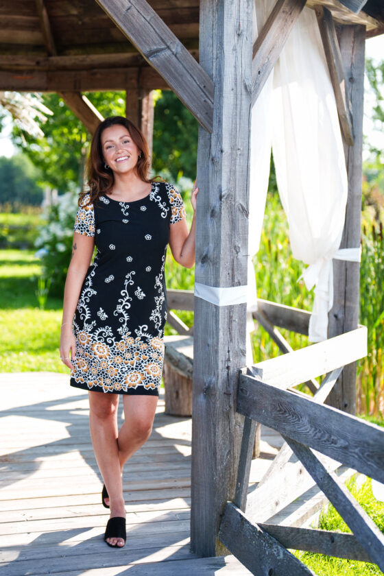 A woman wearing the Classic Dress by Rien ne se Perd in Black and Gold, a semi-fitted black  sheath dress with a gold floral pattern, short sleeves, and an above the knee length. She is standing outside in a pagoda.
