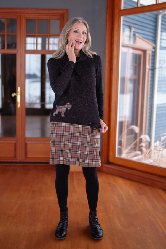 A woman wearing the Gustave Sweater in Charcoal with a plaid Scottish Terrier applique, and a plaid skirt, standing by a picture window. 