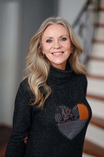 Close up of a woman wearing the Michelangelo Dress by Rien ne se Perd in Grey, a long sleeved turtleneck dress with a heart applique on the front, standing in front of a staircase. 