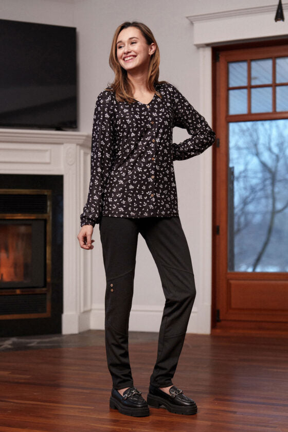 A woman wearing the Monet Blouse by Rien ne se Perd in Black and White Floral, with a V-neck, faux button placket with coconut buttons, loose fit, and long sleeves with gathered cuffs. She is wearing it with black pants nd standing in front of a fireplace. 