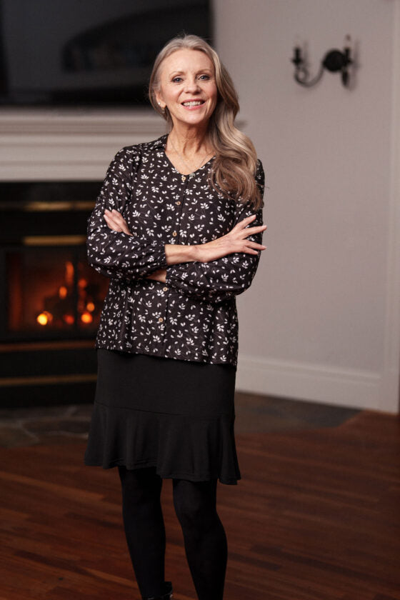 A woman wearing the Monet Blouse by Rien ne se Perd in Black and White Floral, with a V-neck, faux button placket with coconut buttons, loose fit, and long sleeves with gathered cuffs. She is wearing it with a black skirt and standing in front of a fireplace. 