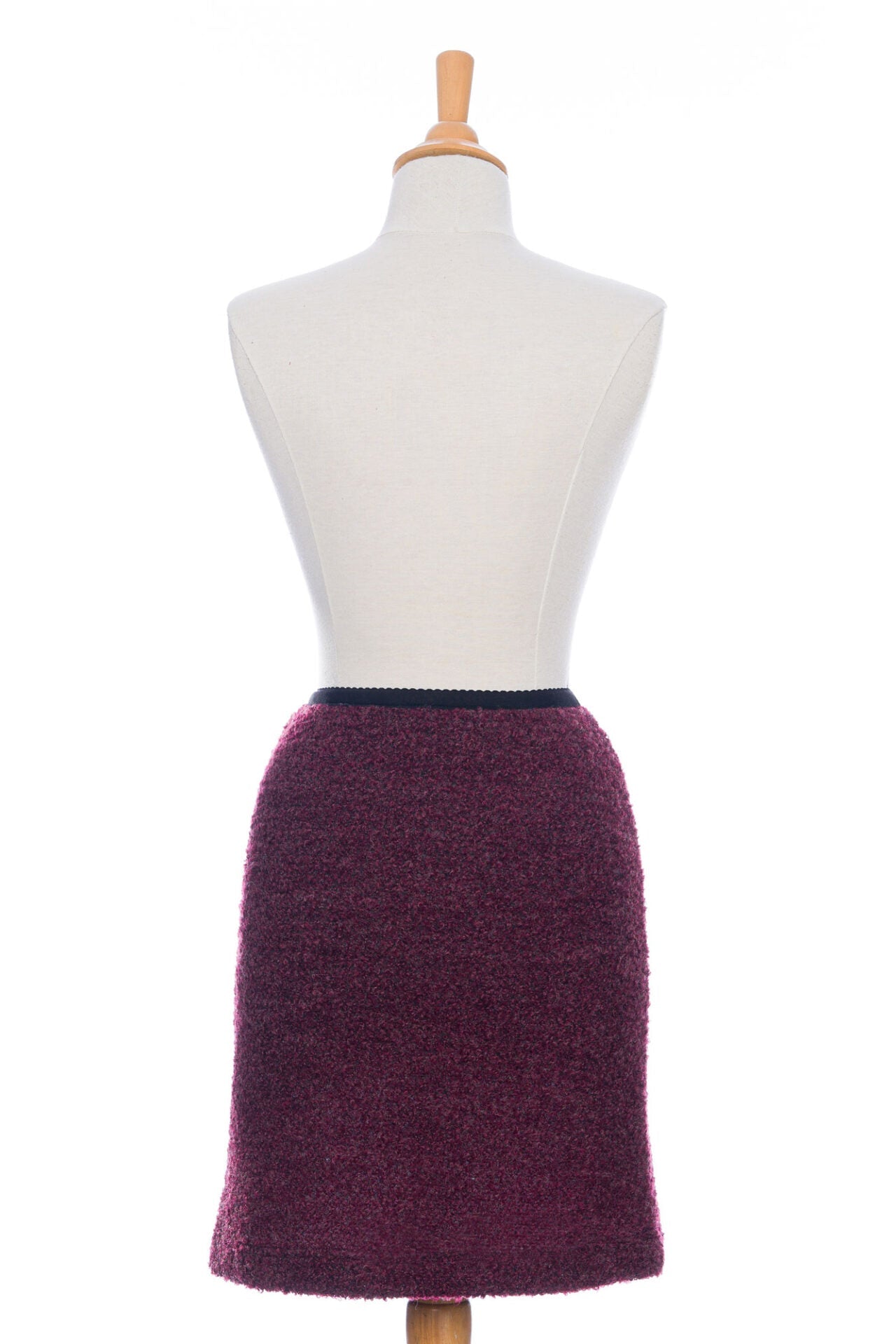 A back view of the Rhododendron Skirt by Rien ne se Perd in Merlot, a straight cut boucle skirt with an elastic waistband and a hem that hits above the knee, is shown on a mannequin in front of a white background.