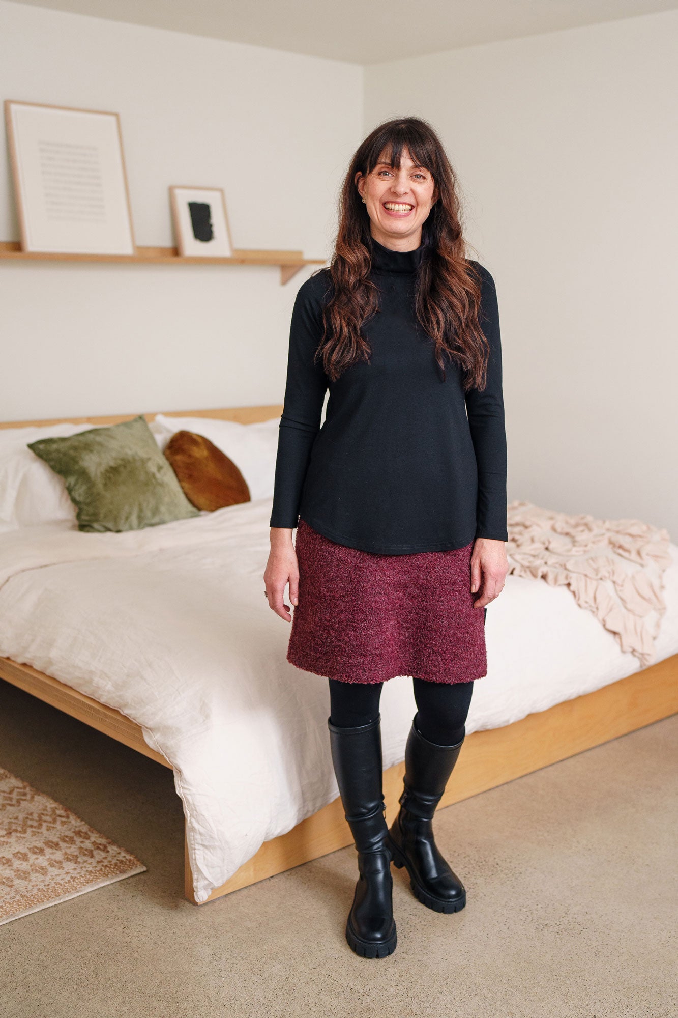 Woman wearing the Rhododendron Skirt by Rien ne se Perd in Merlot, a straight cut boucle skirt with an elastic waistband and a hem that hits above the knee. She is wearing it with a black top and is standing indoors in front of a bed.