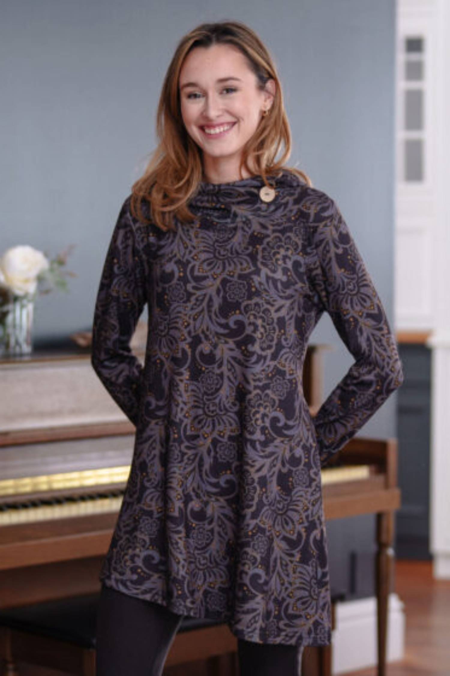 3/4 shot of a woman wearing the Seguin Tunic by Rien ne se Perd in a grey floral pattern with gold accents, standing front of a piano 