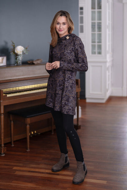 A woman wearing the Seguin Tunic by Rien ne se Perd in a grey floral pattern with gold accents, with black pants, standing front of a piano 