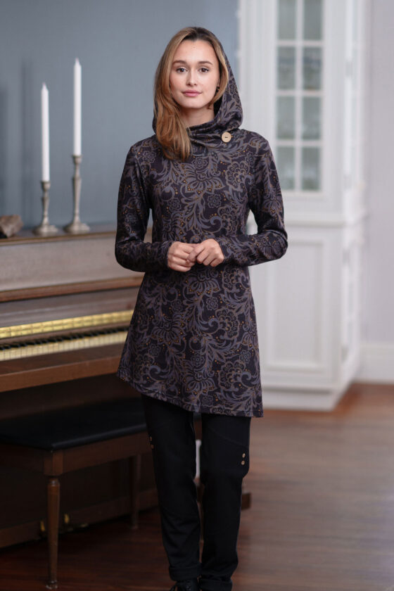 A woman wearing the hooded Seguin Tunic by Rien ne se Perd in a grey floral pattern with gold accents, standing front of a piano 