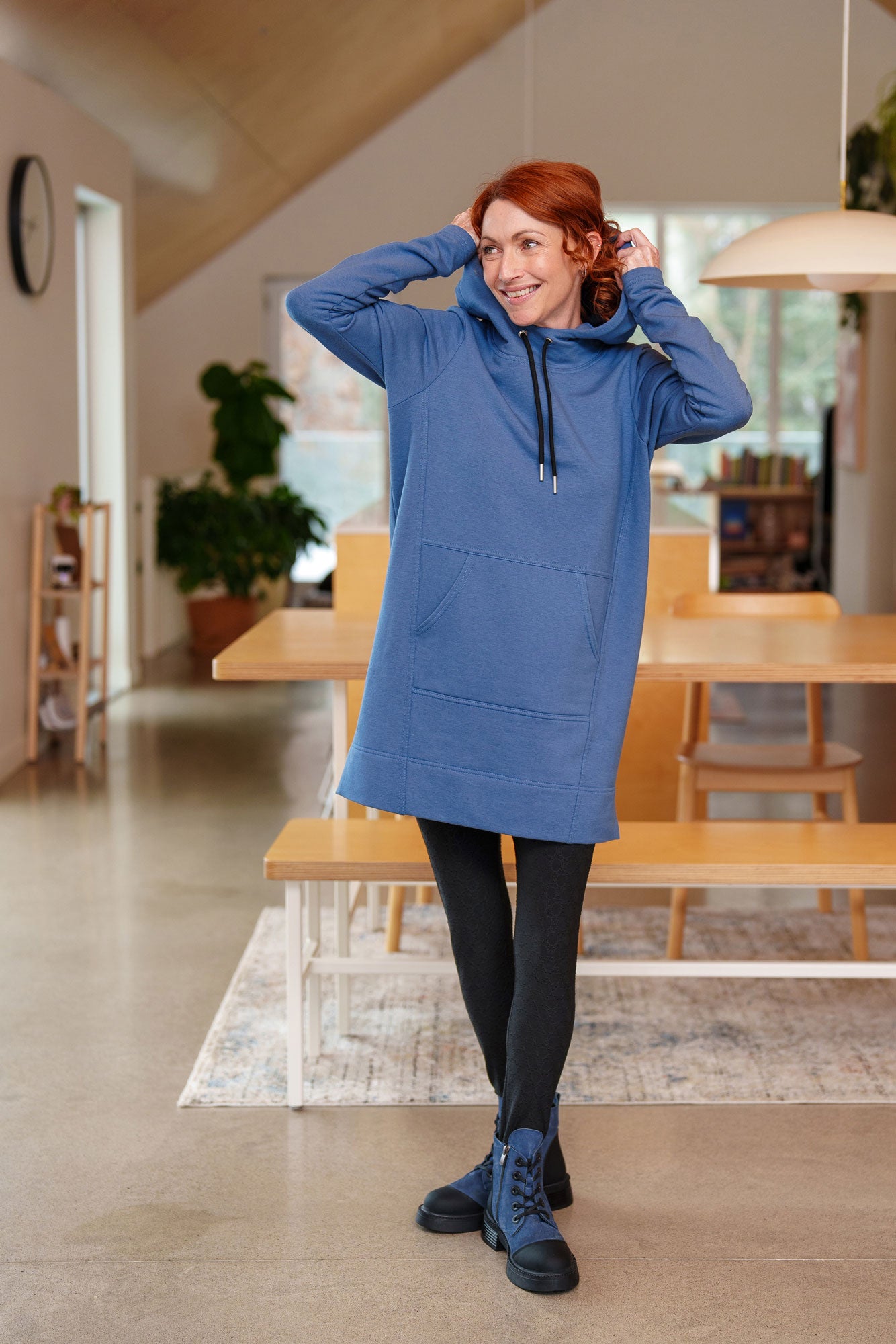 A  woman wearing the Tsuga Leggings by Rien ne se Perd in Black, featuring textured fabric from the mid-thighs down and solid fabric from thigh to waist. She is wearing them with a blue tunic and standing in a kitchen. 
