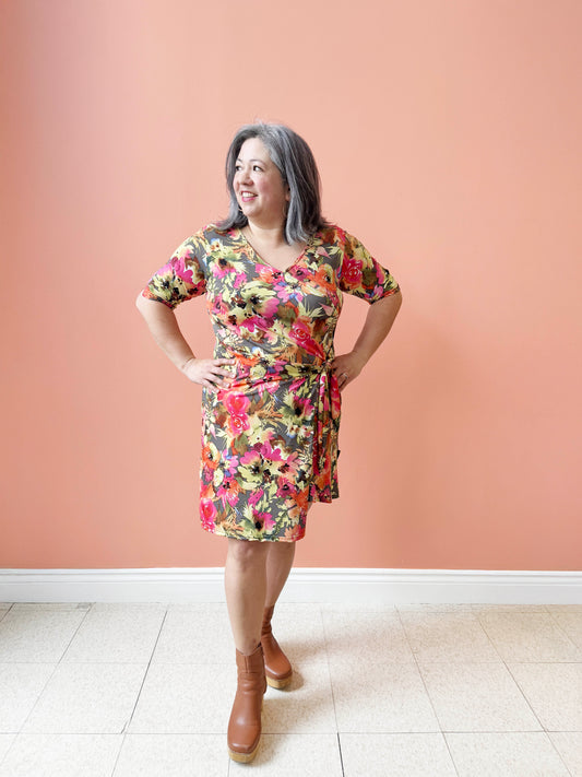 Christina is wearing the Cha Cha Cha Dress by Rien ne se Perd, a faux-wrap dress in a floral pattern with a V-neck, tie at the waist, and knee-length A-line skirt. She is standing in front of a coral background.  