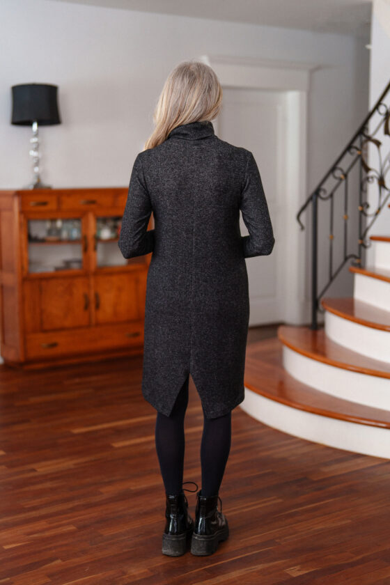 Back view of a woman wearing the Michelangelo Dress by Rien ne se Perd in Grey, a long sleeved turtleneck dress with a heart applique on the front, standing in front of a staircase. 