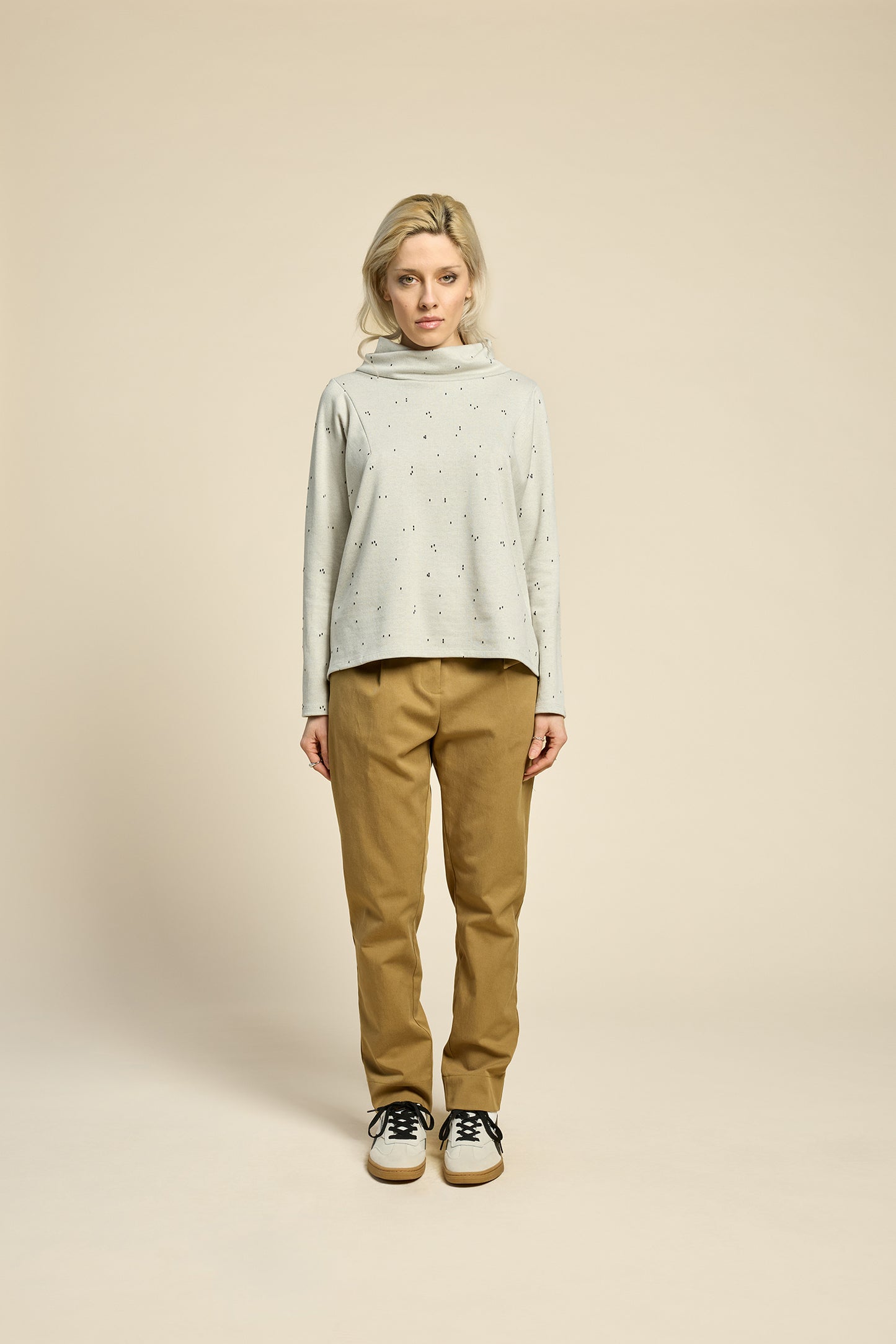 A woman wearing the Reminder Sweater by Cokluch in Natural, with a high twisted neck, darts and the shoulders, long sleeves, and a loose fit. She is wearing it with yellow pants and standing in front of a beige background. 