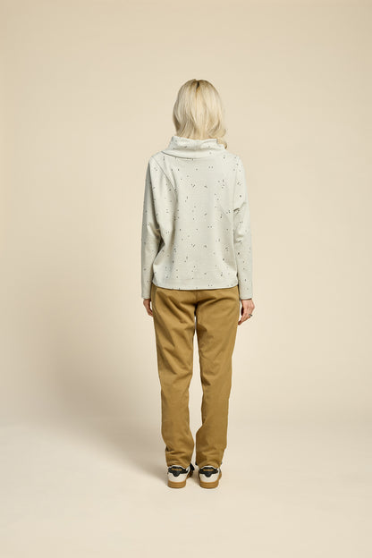 Back view of a woman wearing the Reminder Sweater by Cokluch in Natural, with a high twisted neck, darts and the shoulders, long sleeves, and a loose fit. She is wearing it with yellow pants and standing in front of a beige background. 