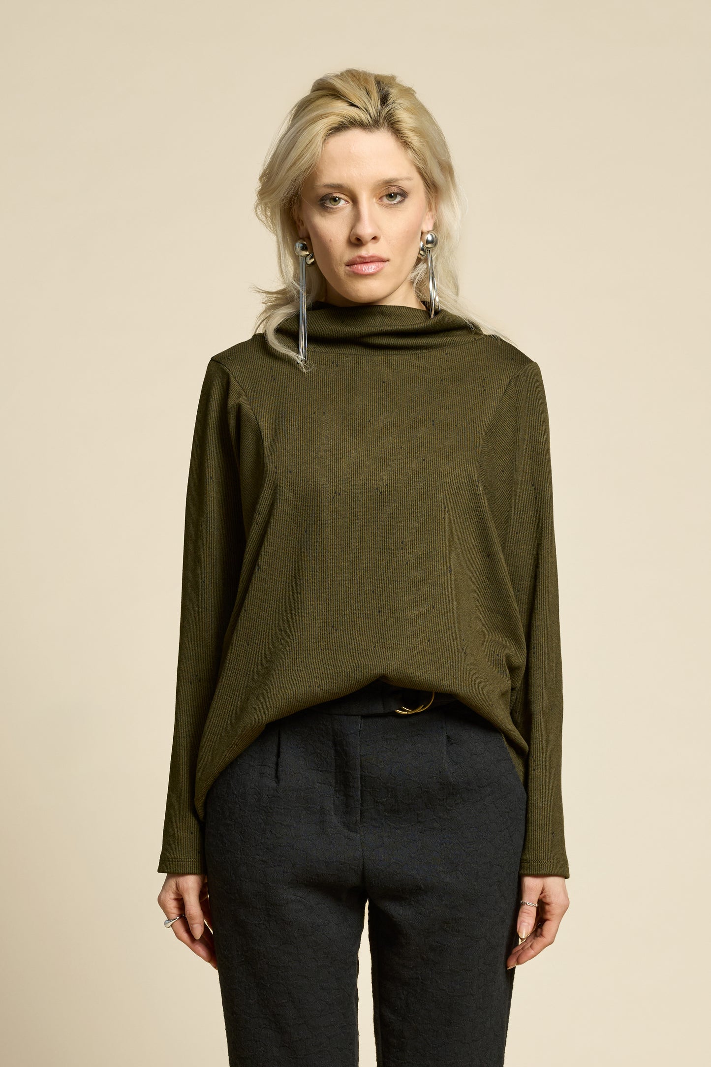 Waist up view of a woman wearing the Reminder Sweater by Cokluch in Olive, with a high twisted neck, darts and the shoulders, long sleeves, and a loose fit. She is wearing it with black pants and standing in front of a beige background. 