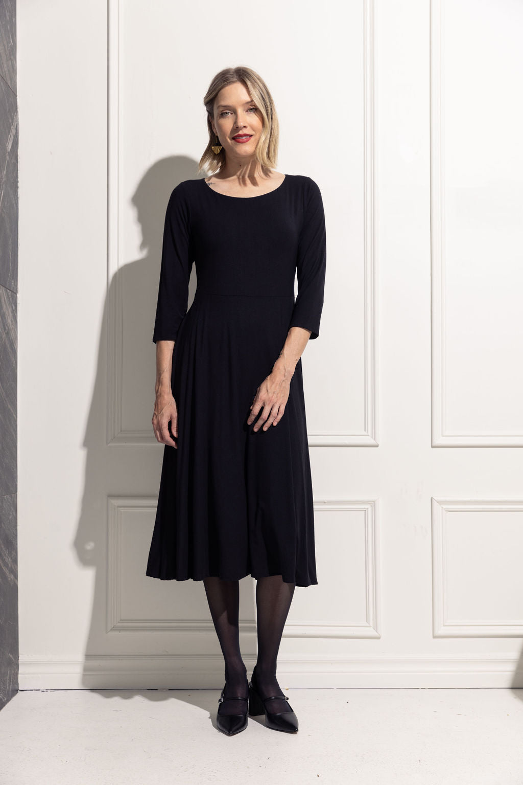 A woman wearing the Diamond Dress by Cherry Bobin in Black, featuring a boat neck, 3/4 sleeves, a fit and flare shape, and a tea-length skirt in bamboo rayon. She is standing. in front of a white wall. 