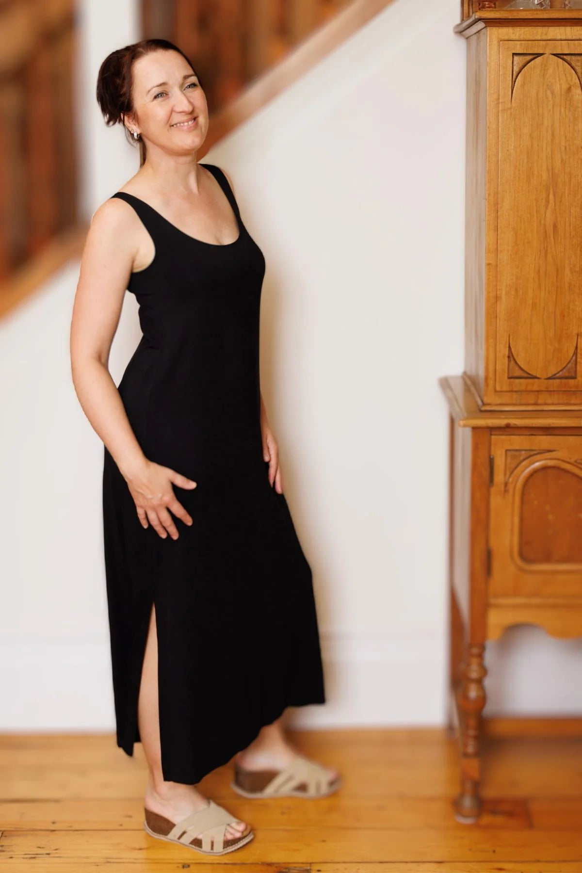 A woman wearing the Ruby Dress by Moovment in Black, a long camisole dress with side slits. She is standing against a white wall beside a wooden cupboard. 