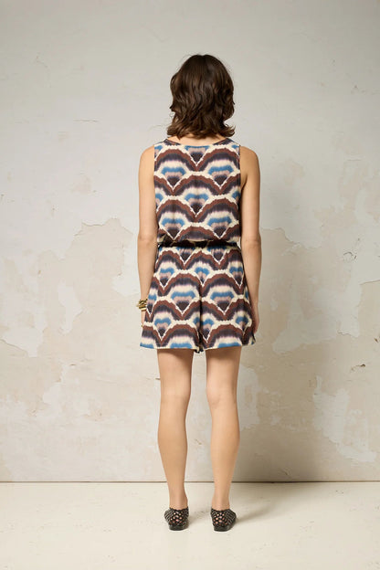 A woman wearing the Atman Shorts by Cokluch in Feather print, mid-thigh length shorts with a high elastic waist and an attached ring belt. She is wearing them with a matching top and standing in front of a plain background. 
