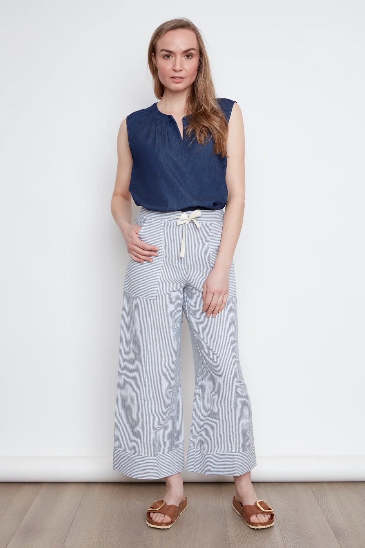A woman wearing the Sabrina Top by Julei in Denim, with raglan cap sleeves, a notched v-neck with pleat details, and a pleated back yoke. She is wearing it with striped pants and standing against a white background