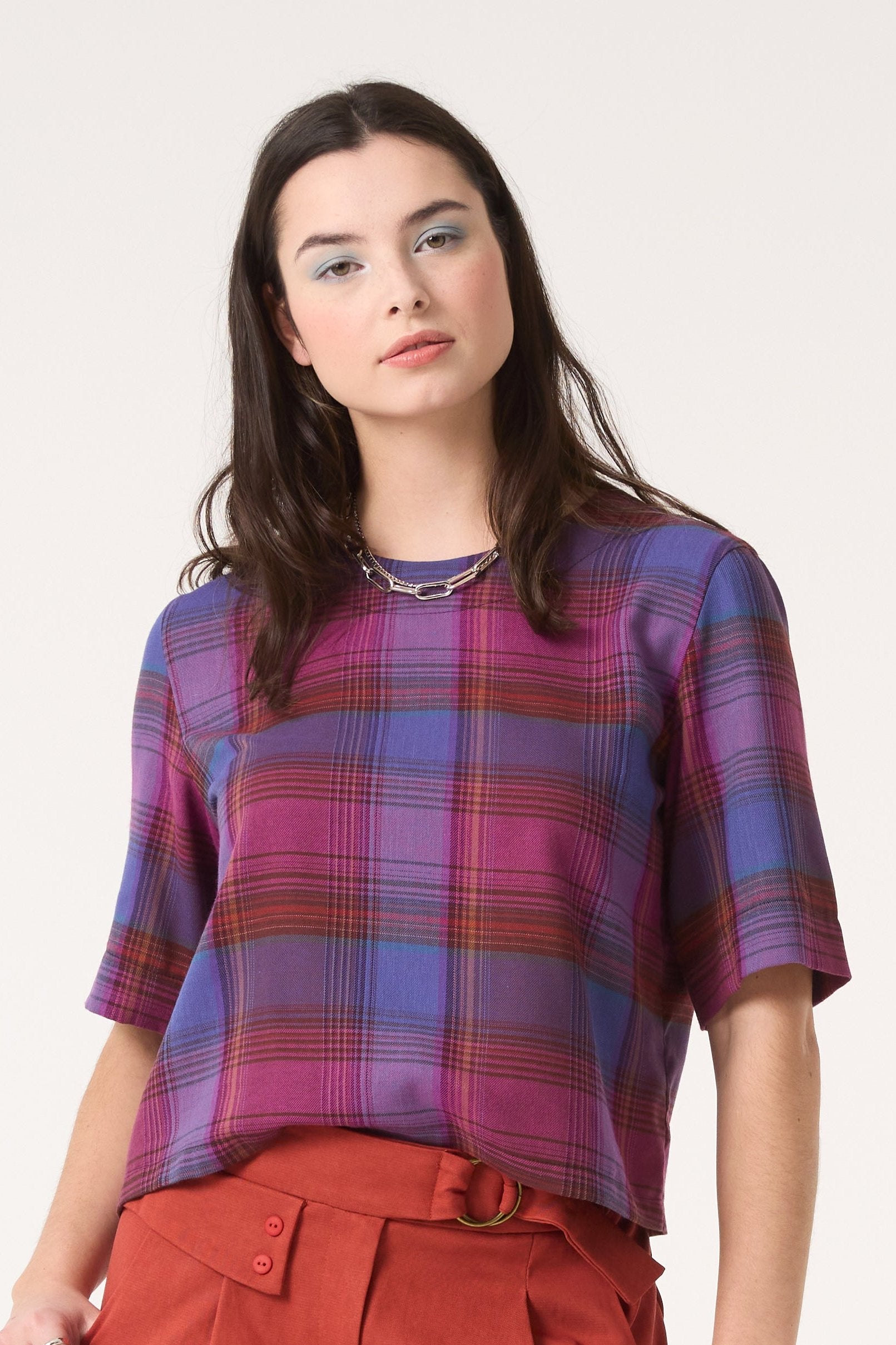 Semi close-up of a woman wearing the Sideral Top by Cokluch in Purple Plaid with orange pants, standing in front of white background 