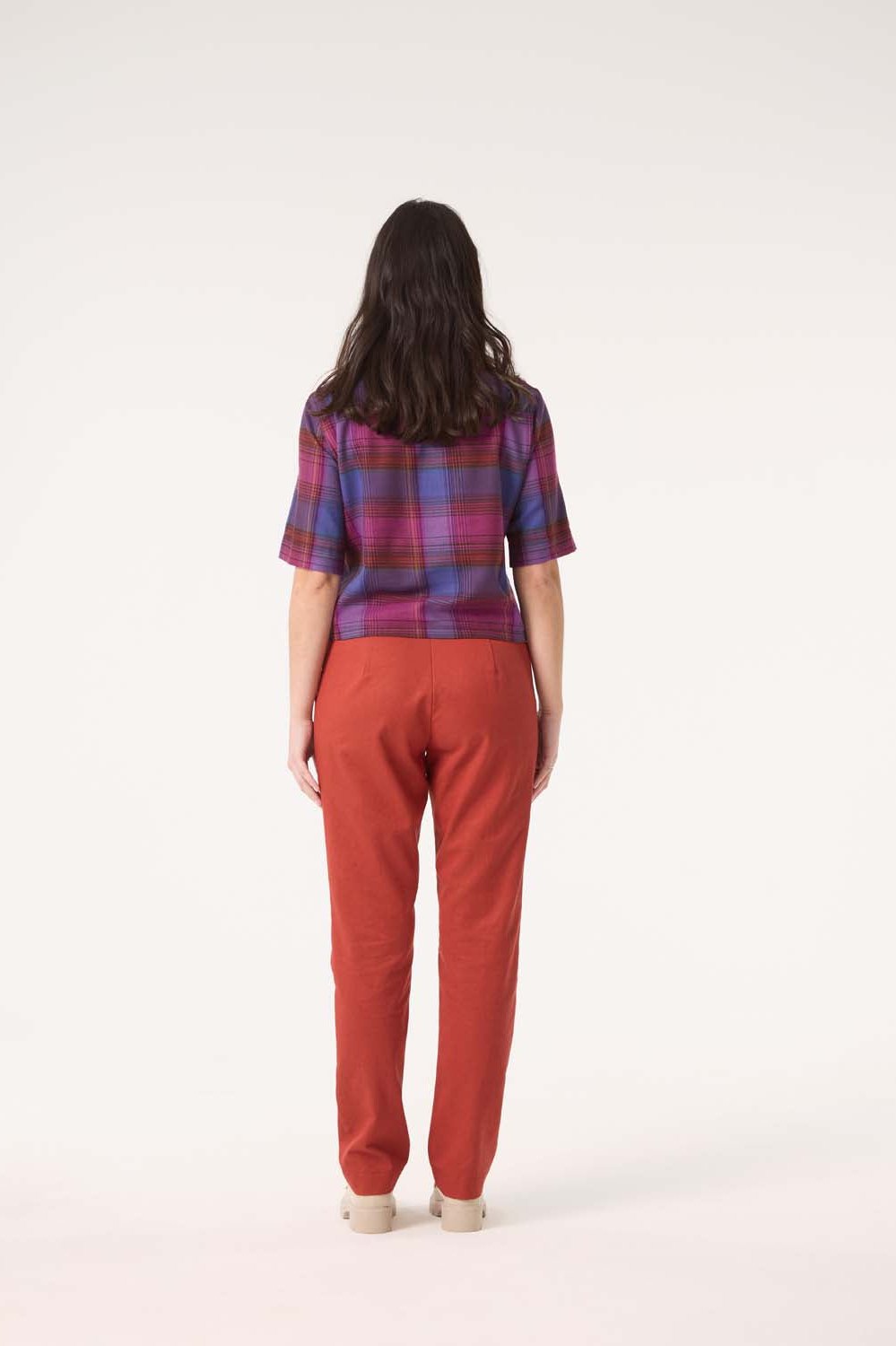 Back view of a woman wearing the Sideral Top by Cokluch in Purple Plaid with orange pants, standing in front of white background 