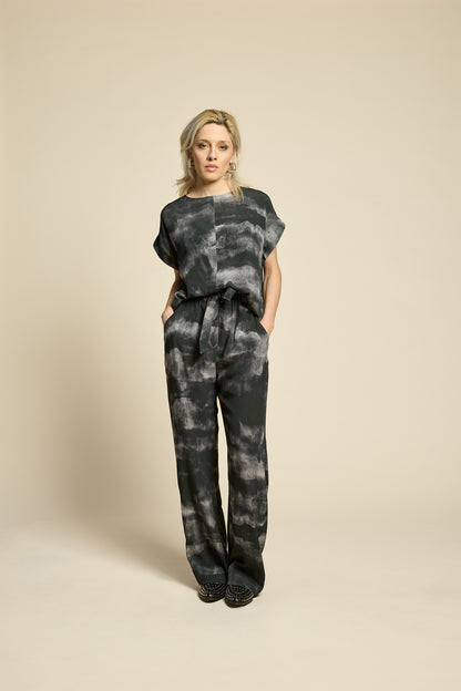 A woman wearing the Silence Top by Cokluch in Mist Black, with a round neck, short Magyar sleeves, and a loose fit. She is wearing it with matching pants and standing in front of a beige background. 