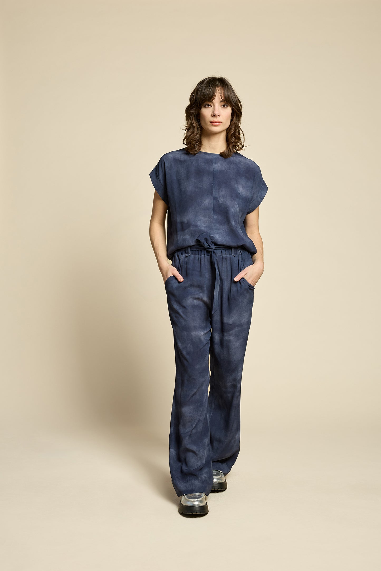 A woman wearing the Silence Top by Cokluch in Mist Twilight, with a round neck, short Magyar sleeves, and a loose fit. She is wearing it with matching pants and standing in front of a beige background. 