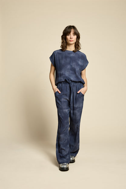 A woman wearing the Silence Top by Cokluch in Mist Twilight, with a round neck, short Magyar sleeves, and a loose fit. She is wearing it with matching pants and standing in front of a beige background. 