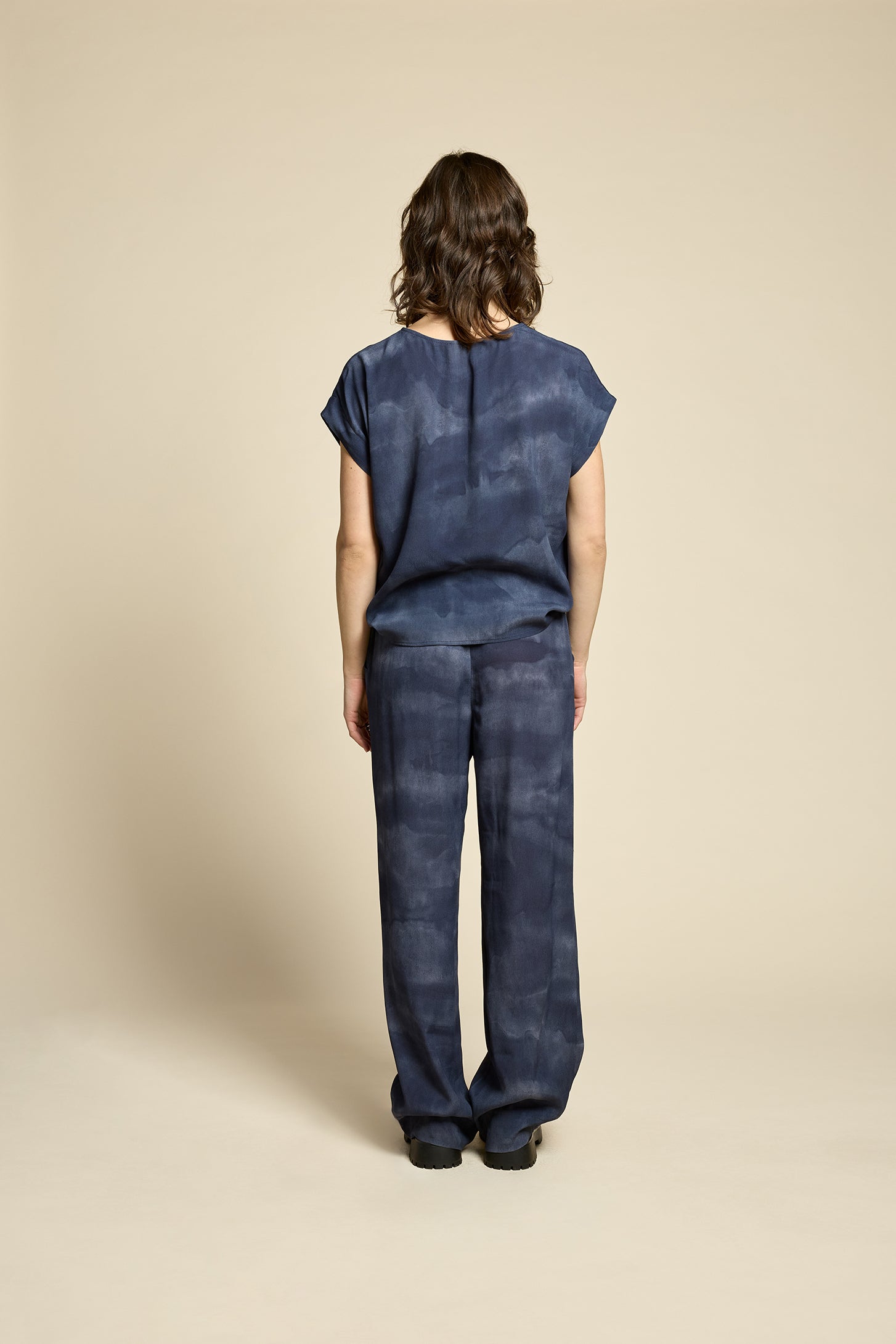 Back view of a woman wearing the Silence Top by Cokluch in Mist Twilight, with a round neck, short Magyar sleeves, and a loose fit. She is wearing it with matching pants and standing in front of a beige background. 