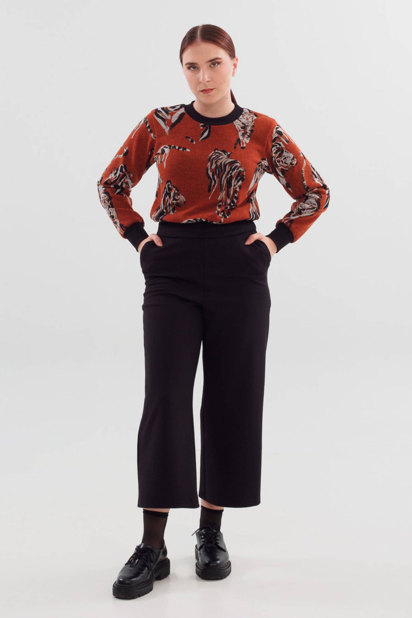 A woman wearing A24 Pants by Slak in Black, featuring a pull-on waist and wide cropped legs, with the Wanda Sweater in Tiger, standing in front of a white background 