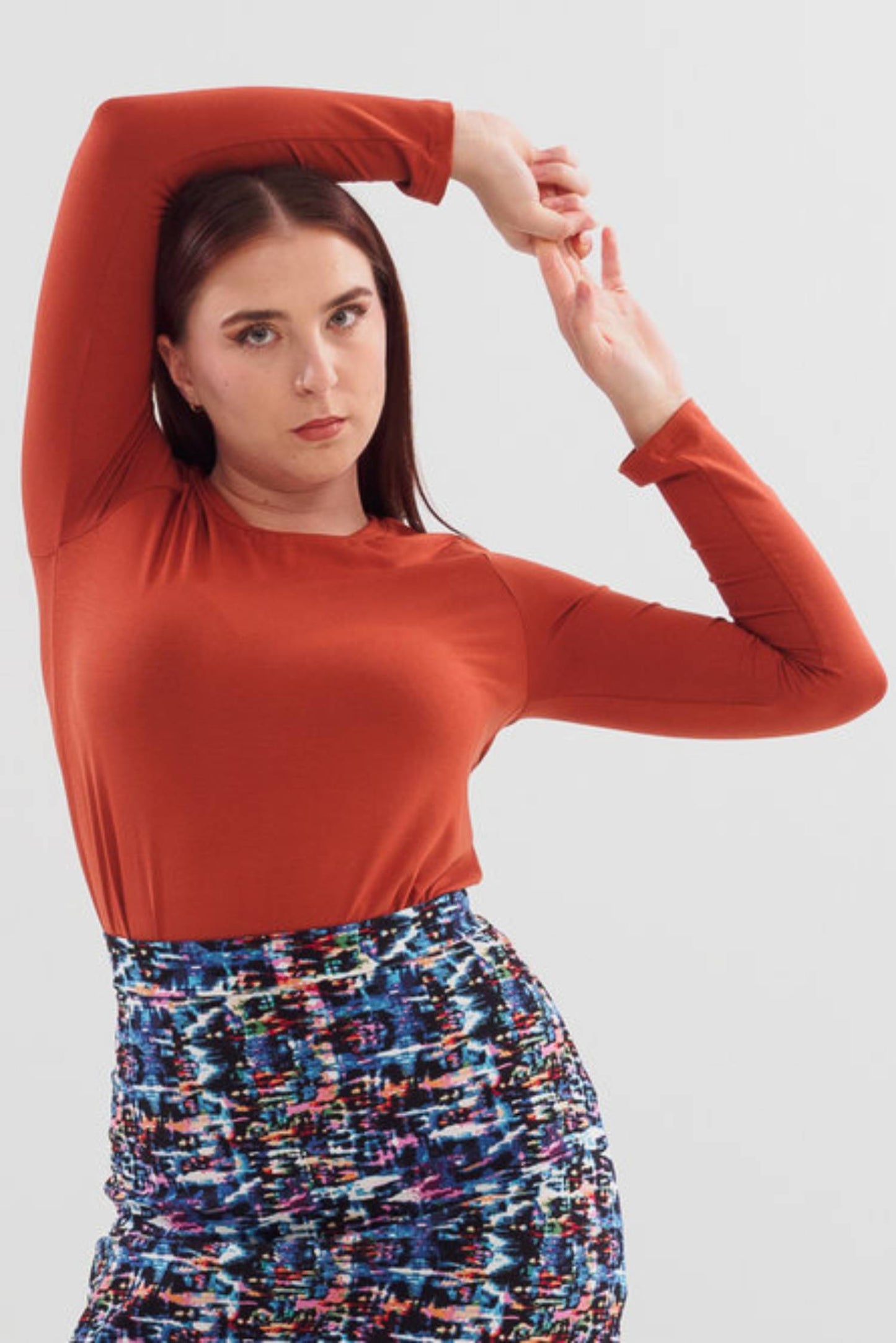 A woman wearing the A24 Top by Slak in Caramel, featuring long sleeves and a round neck, with the Eve Skirt. She is standing in front of white background. 