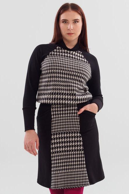 A woman wearing the Nena Sweater by Slak in Black and White houndstooth, with contrasting black raglan sleeves, and ribbing at hem, cuffs and cross-over neckline. She is wearing it with a houndstooth skirt and standing in front of a white background 